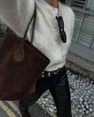 Woman wearing white Sezane cardigan, black jeans, and brown suede bag taking outfit selfie from above.