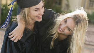 Two young women in graduation gowns, smiling with arms around each other.