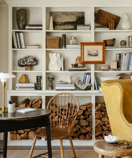 White bookcase styled with vintage decor, books, vases and paintings and logs beneath, a round wooden table with a spindle chair and desk lamp, and a yellow wingback leather armchair beside a rustic wooden stool