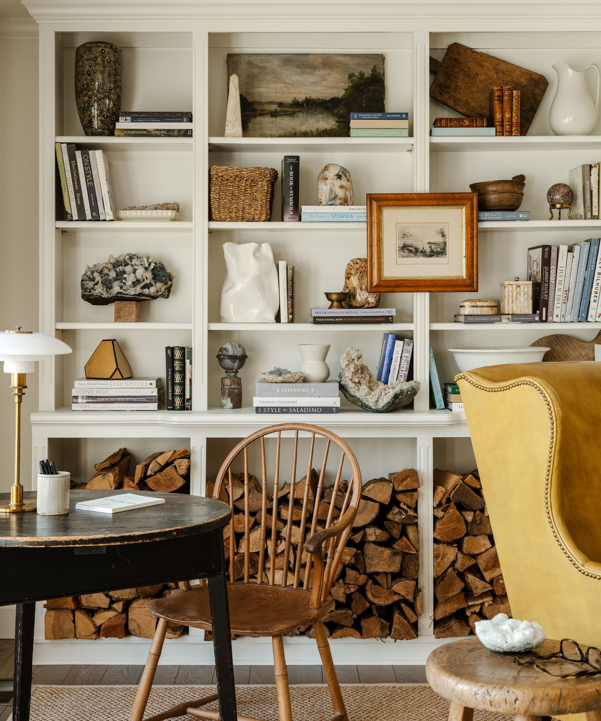 White bookcase styled with vintage decor, books, vases and paintings and logs beneath, a round wooden table with a spindle chair and desk lamp, and a yellow wingback leather armchair beside a rustic wooden stool