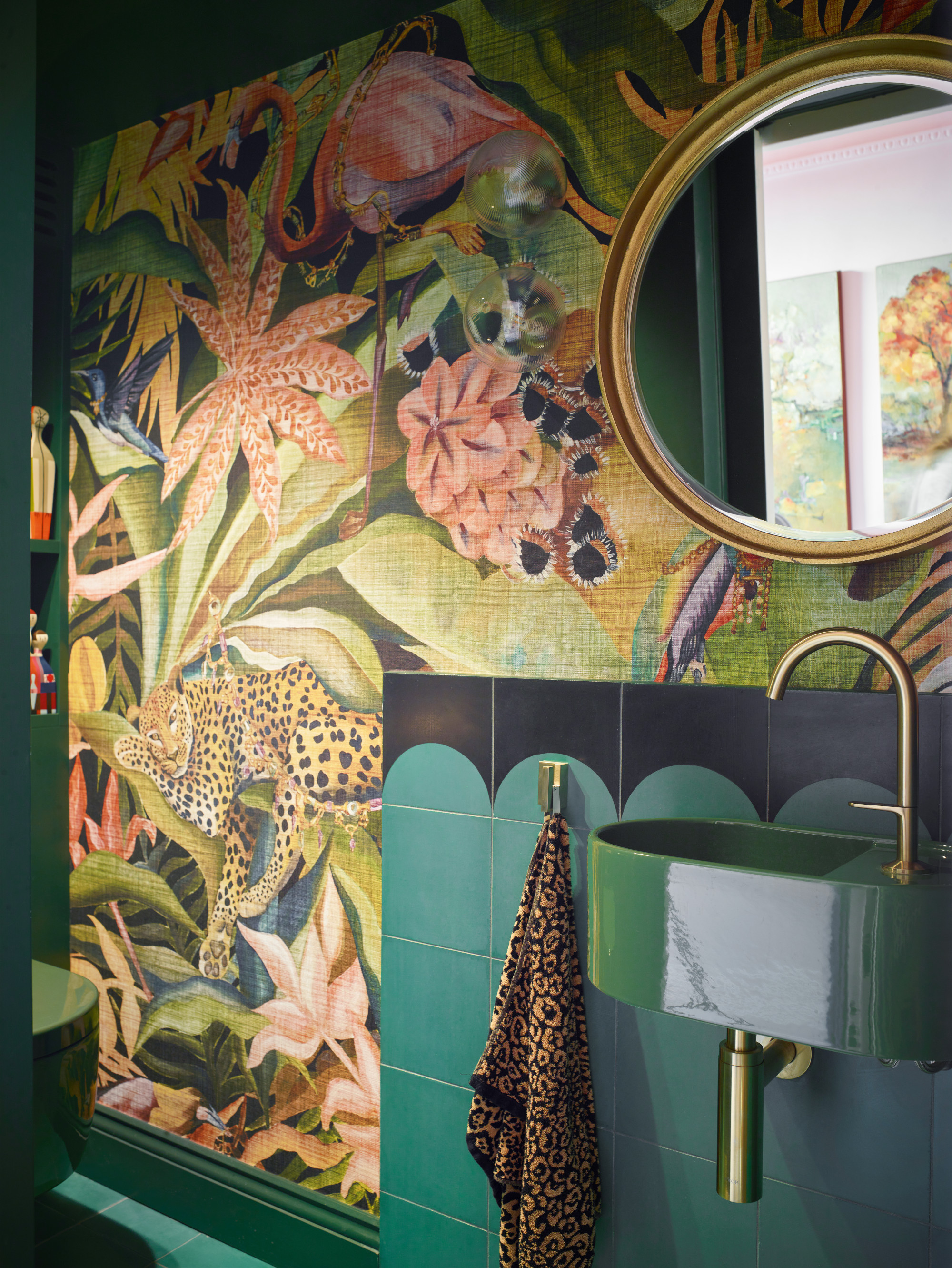 Bathroom with jungle wallpaper, green tiles, green sink and round brass mirror with leopard print towel