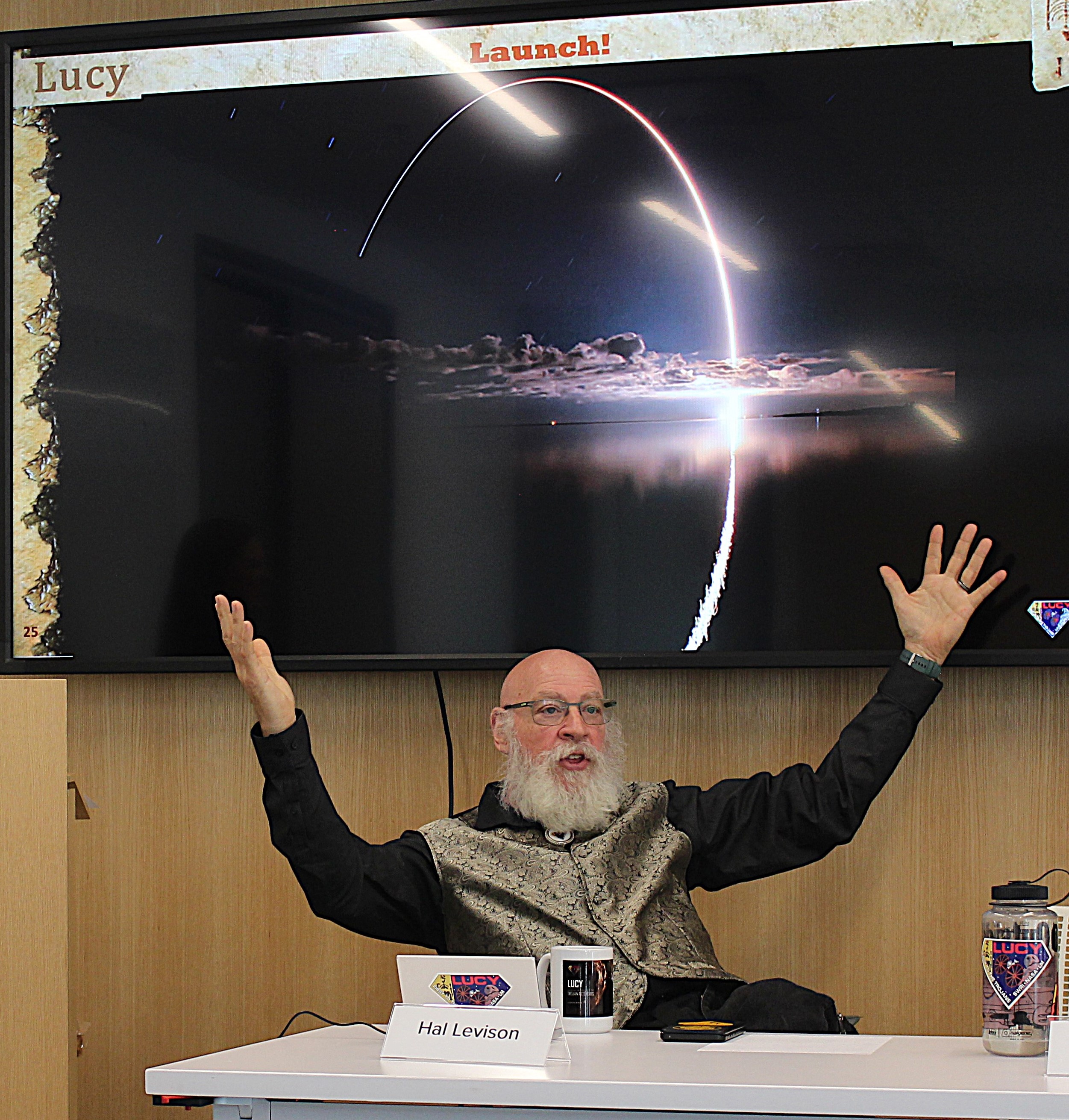 a bald man in a button-down shirt and bolo tie stands in front of a television screen with a photo of an irregularly-shaped rock on it