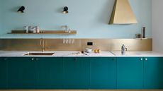 A baby blue colored kitchen with teal kitchen cabinets and marble worktops, gold backsplash and tiled floors.