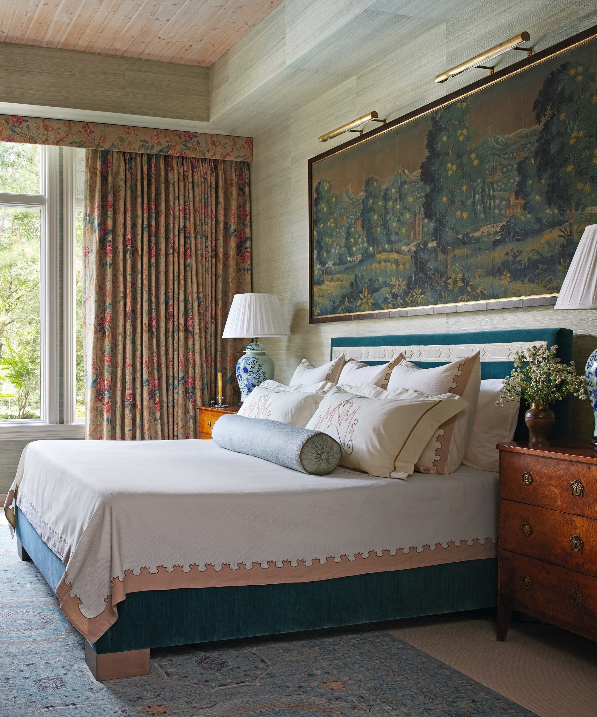 A sophisticated bedroom with seafoam green grasscloth walls, a large landscape tapestry above a teal velvet bed, and floral patterned curtains that match the traditional wooden nightstands