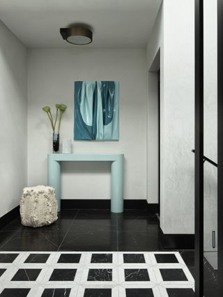 Image of a white and black entryway with a white and black checkered floor that turns into solid black. There is a light cyan console table with an abstract, 3D painting in a cyan color hanging above it. On the table is a vase with long green flowers