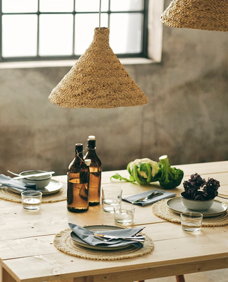 A dining table with woven placemats and a woven pendant lampshade overhead