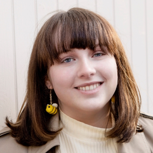 Image of a woman with brown hair and bangs wearing a light yellow turtleneck, a khaki trench coat, and bumble bee earrings.