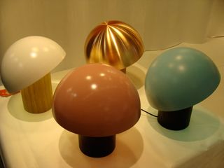 Four mushroom-like designed table lights in pink, green, white and gold photographed on a white surface