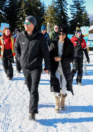 Prince Harry walking in the snow holding hands with Meghan Markle, both wearing black winter coats and hats