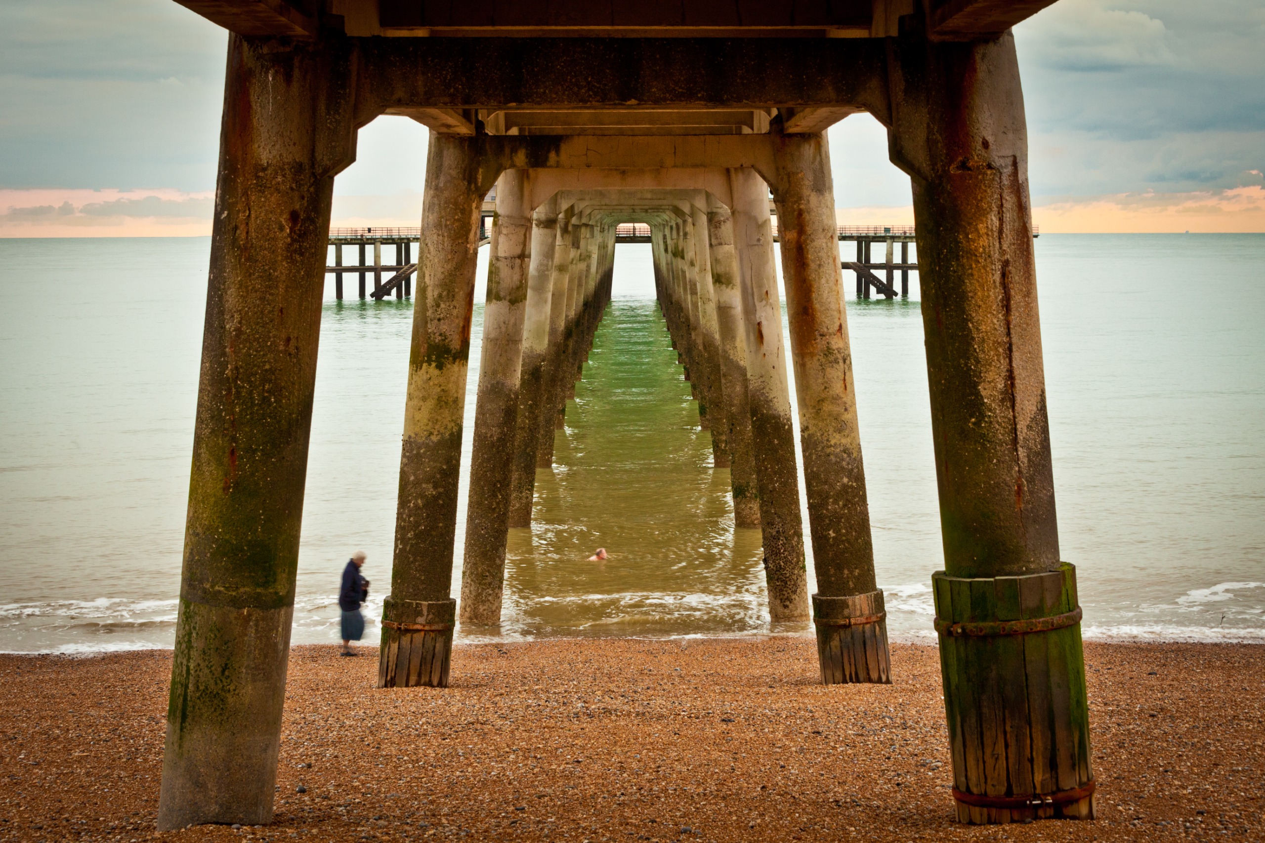 The underneath of a wooden pier stretching out into the sea. A single woman stands next to one of the tall struts