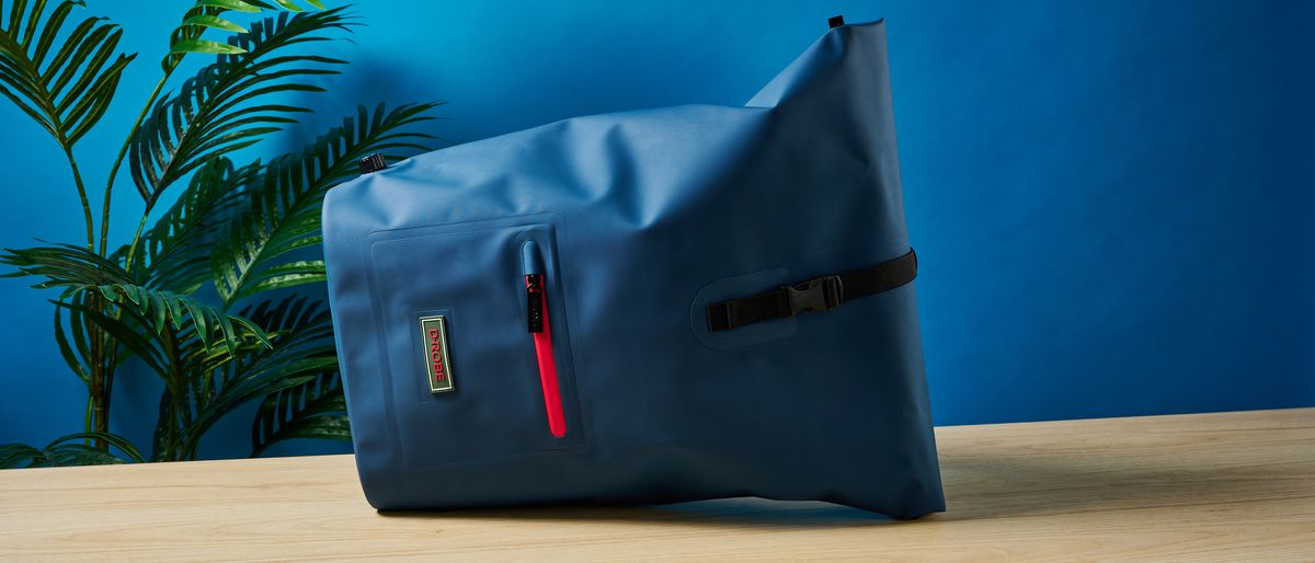 A photo of the D-Robe Roll-Top backpack on its side, on a table with a blue wall and green plant in the background