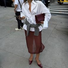 style influencer Caroline Lin poses in Chicago holding her cell phone wearing a chic outfit with a white oversize button-down shirt, burgundy clutch bag, a gray sweater tied around the waist, a sheer burgundy skirt, and patent burgundy slingback heels