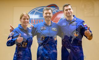 three smiling astronauts in blue flight suits give a thumbs up.