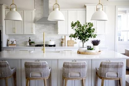 white kitchen with white countertops, pearlescent backsplash tiles, brass faucet, white pendants, beige woven bar stools