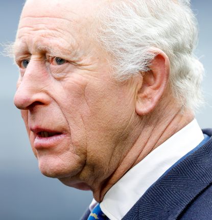 A headshot of King Charles wearing a blue suit and white shirt turned to the left and looking serious