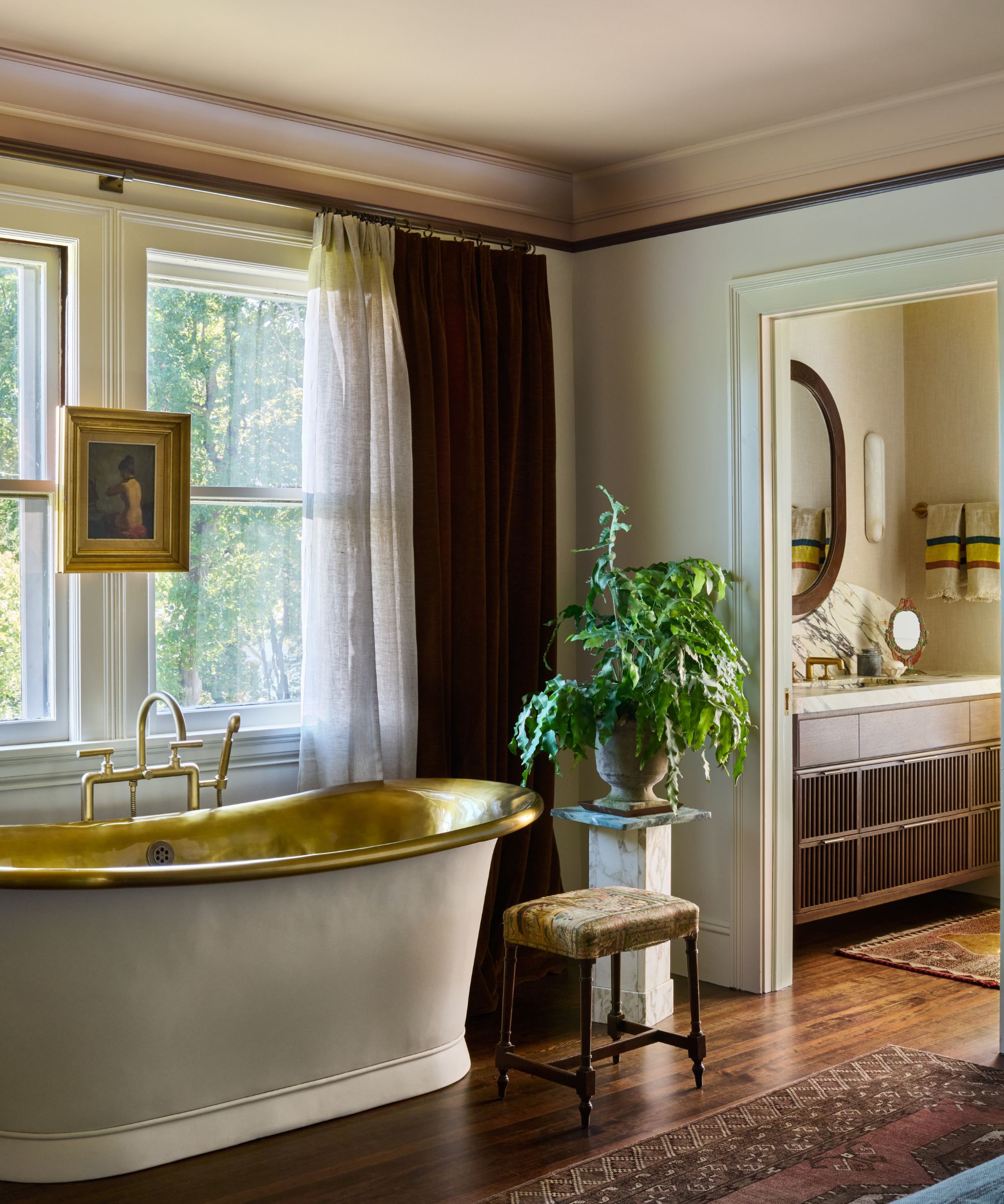Bath positioned next to a window with a piece of art hung in front of the window. Wooden flooring, stool and plant next to bath, view into bathroom with vanity and mirror