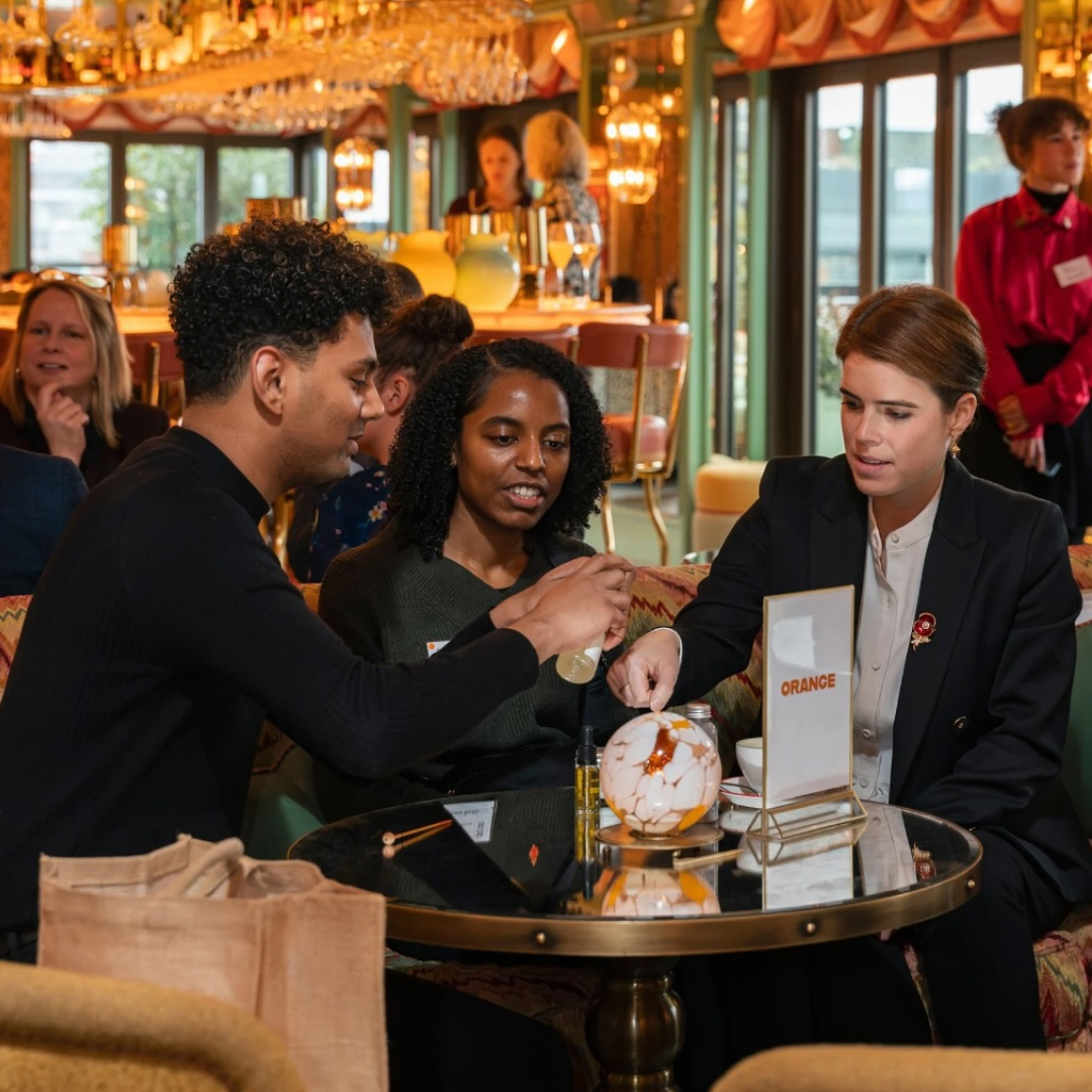 Princess Eugenie sitting at a table with a man and woman trying a product on her hand