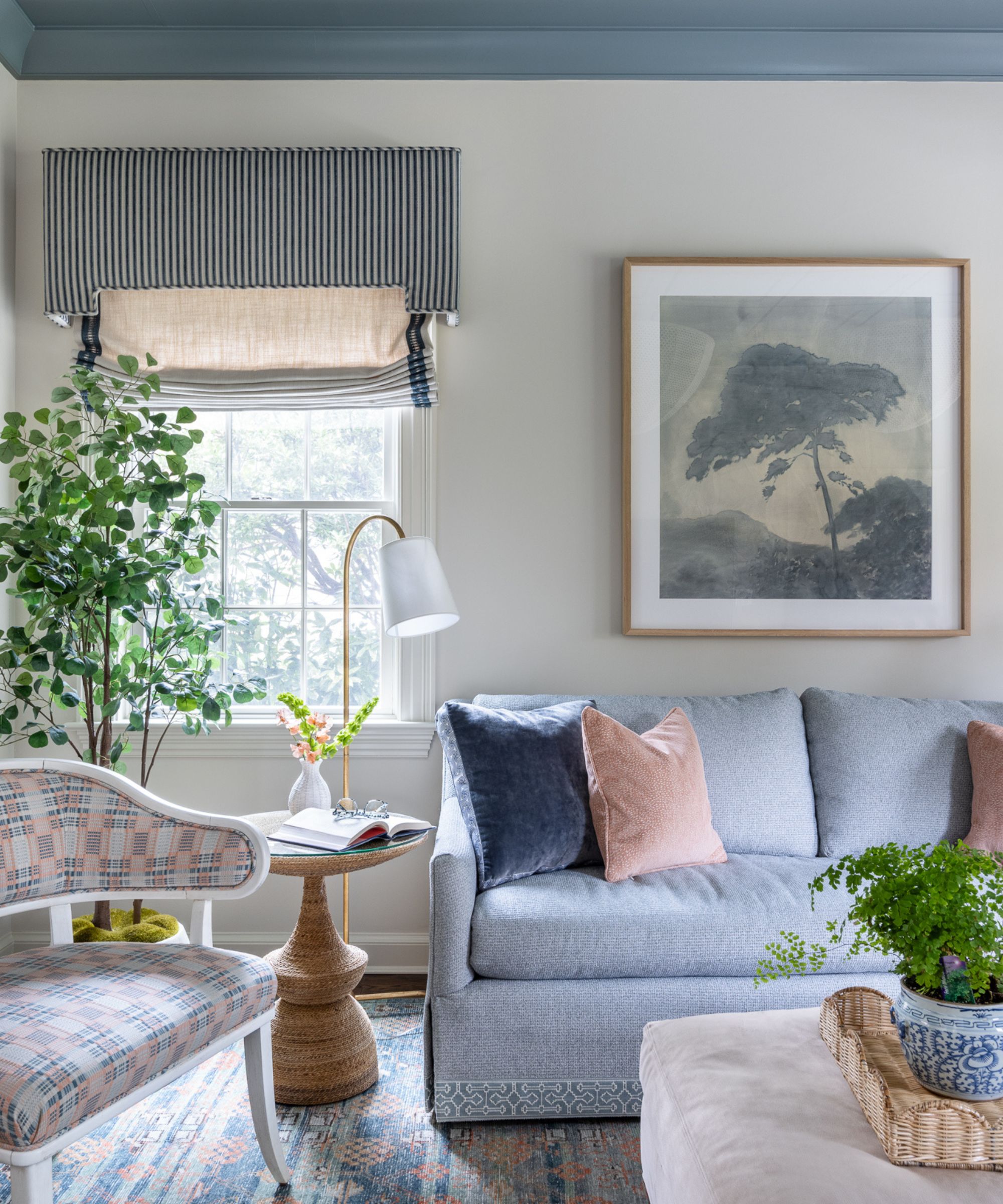 A transitional living room with off-white walls and a mid-blue ceiling, a powder blue sofa with dark blue and pink cushions, a dark blue Roman blind, a soft blue artwork, and a patterned armchair.