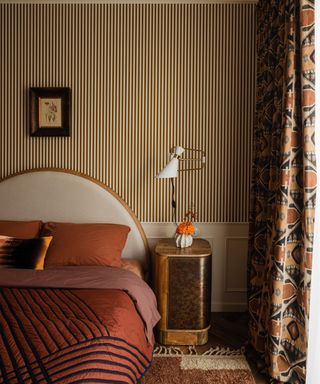 Art deco/ nouveau style bedroom with striped wallpaper, curved headboard and orange bedding, geometric curtains and burl wood and gold bedside table