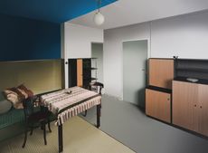 A room with with 2 different colours. The left side of the room has blue wall and ceiling with a table and 2 chairs and wooden floor. The right of the room has white walls and ceiling with a grey door and grey floor. brown and black standing cabinets on the righ of the room. 
