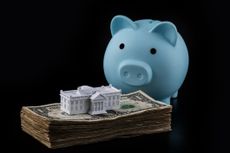 blue piggy bank next to a stack of dollar bills with a miniature white house on top