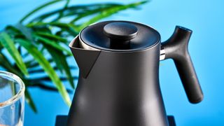 the fellow Corvo EKG kettle in matte black photographed against a blue background with a glass mug and black and white table