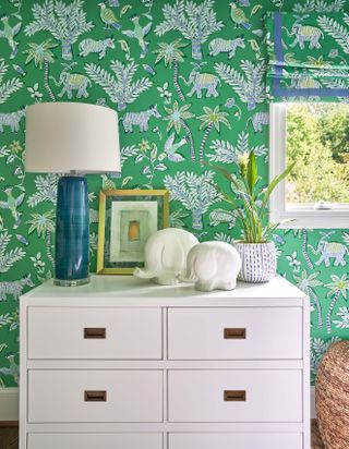 A green wallpapered background with a white chest of drawers styled in front of it