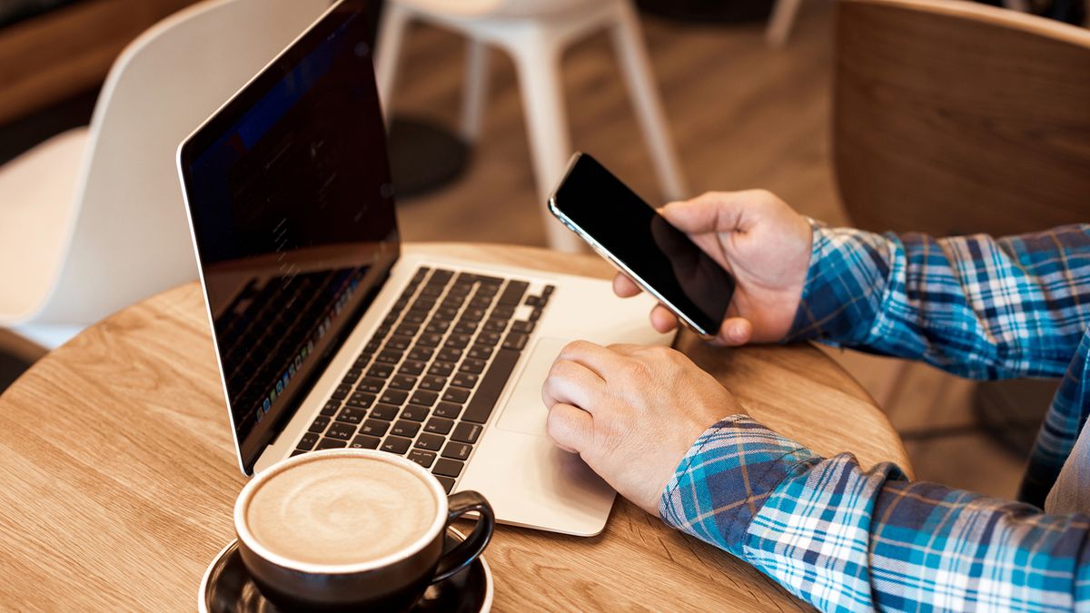 Man using phone and macbook simultaneously in a coffee shop with mug of coffee on table.