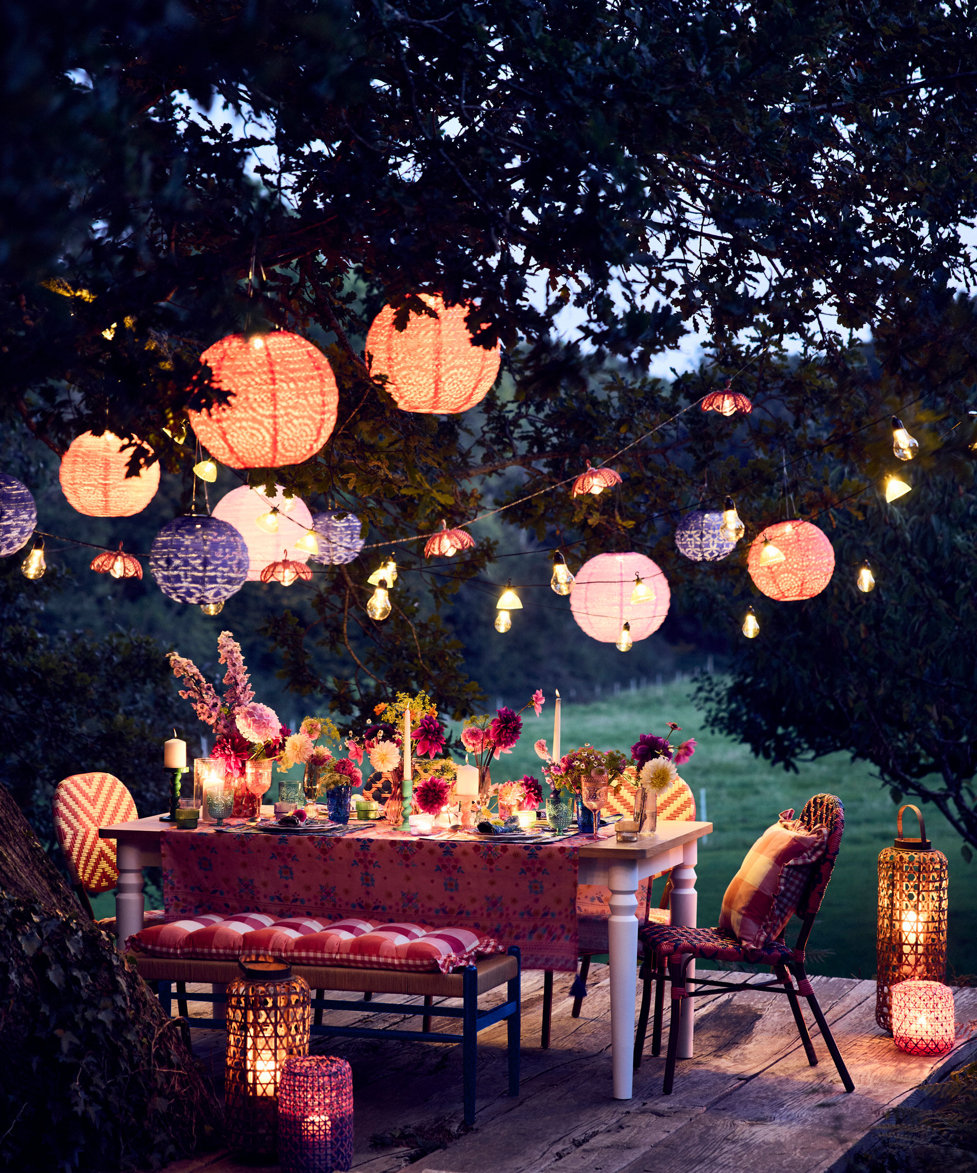 tablescaped garden table and chairs with flowers and candles on a deck, with lanterns hanging from the tree above