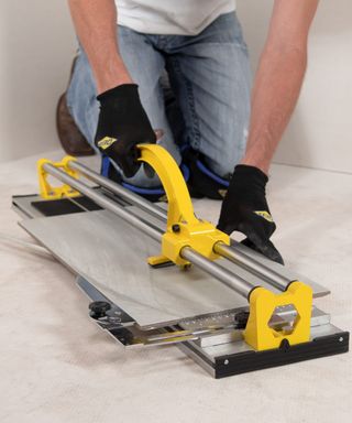 A man wearing utility gloves, white t-shirt and blue denim pants using a yellow QEP 24-inch manual tile cutter