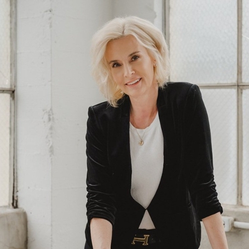Image of a woman in a black blazer and white shirt standing in a white room.