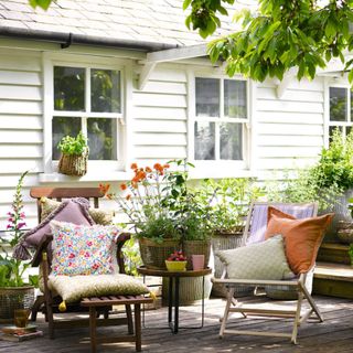 Garden chairs with cushions on a shady deck with pots of plants and flowers and a white wooden clad house behind.