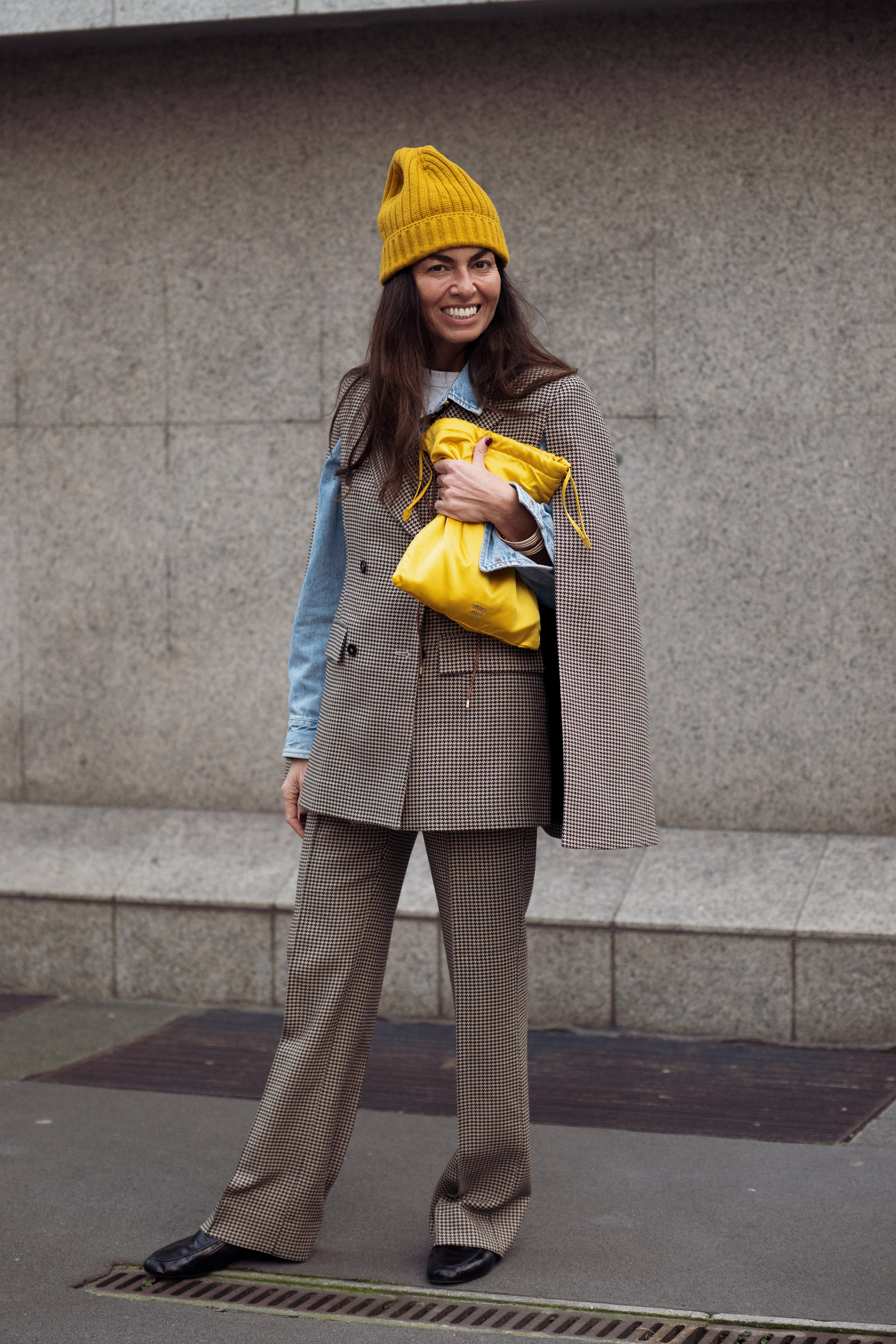 MILAN, ITALY - FEBRUARY 24: A guest wears a brown and black micro-check tailored blazer with matching wide-leg trousers, layered over a light blue denim shirt and styled with a mustard ribbed knit beanie and black leather shoes, carrying a yellow satin drawstring pouch outside Alberta Ferretti presentation during the Milan Fashion Week - Womenswear Fall/Winter 2026/2027 on February 24, 2026 in Milan, Italy. (Photo by Raimonda Kulikauskiene/Getty Images)