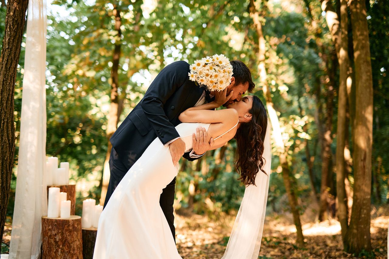 Groom holding a bouquet, bending the bride over for a kiss during a romantic forest wedding ceremony adorned with candles and flowing white curtains, surrounded by nature's beauty