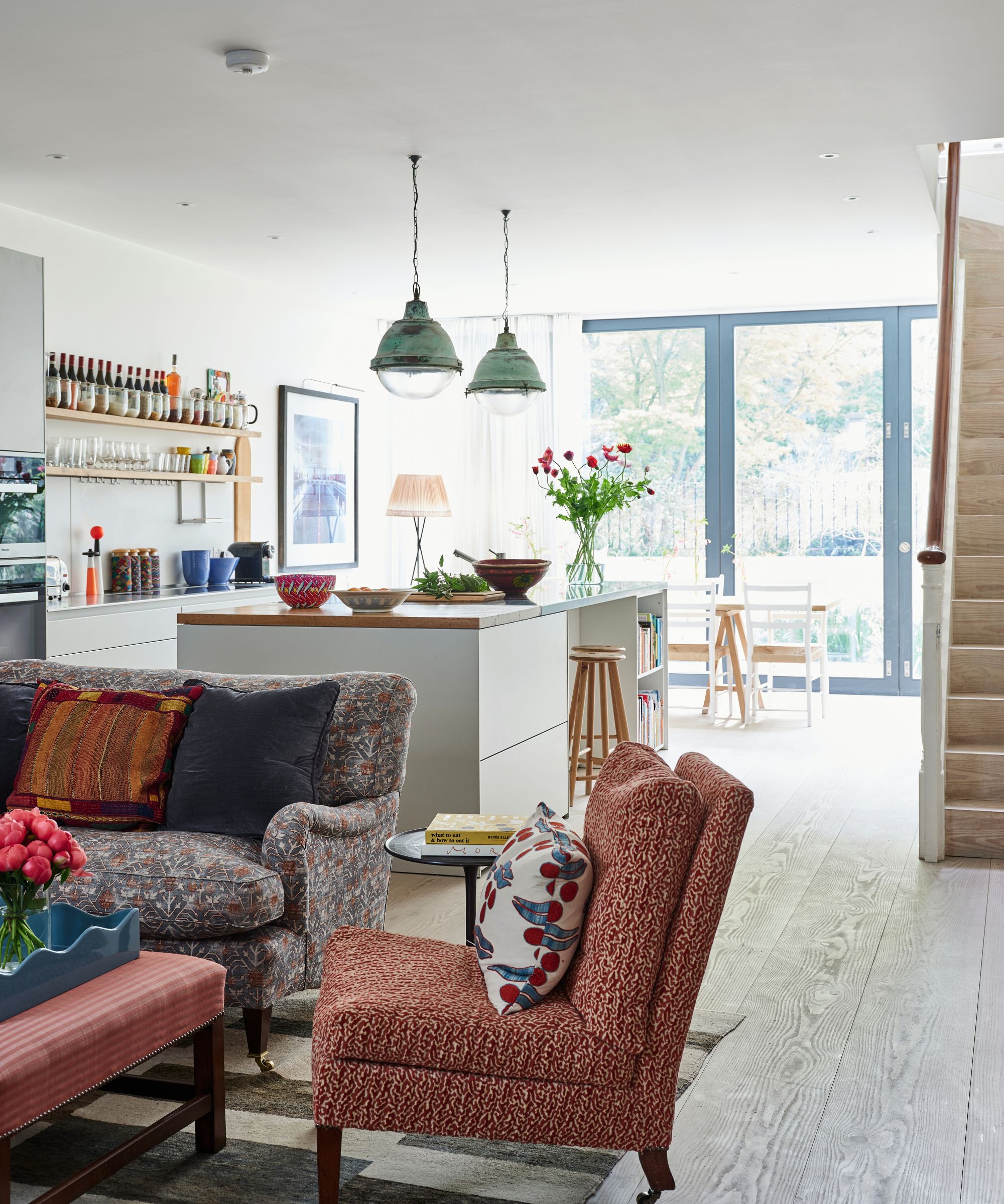 an open plan living room and kitchen with traditional furniture, a large kitchen island with pendants hanging from the ceiling, and a wall of glass doors in the background
