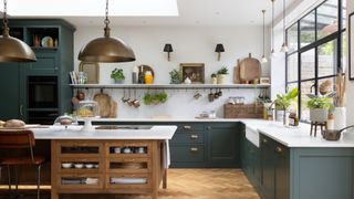 Dark green shaker kitchen with brass pendant lights, open shelving, and a wooden island, parquet floor.
