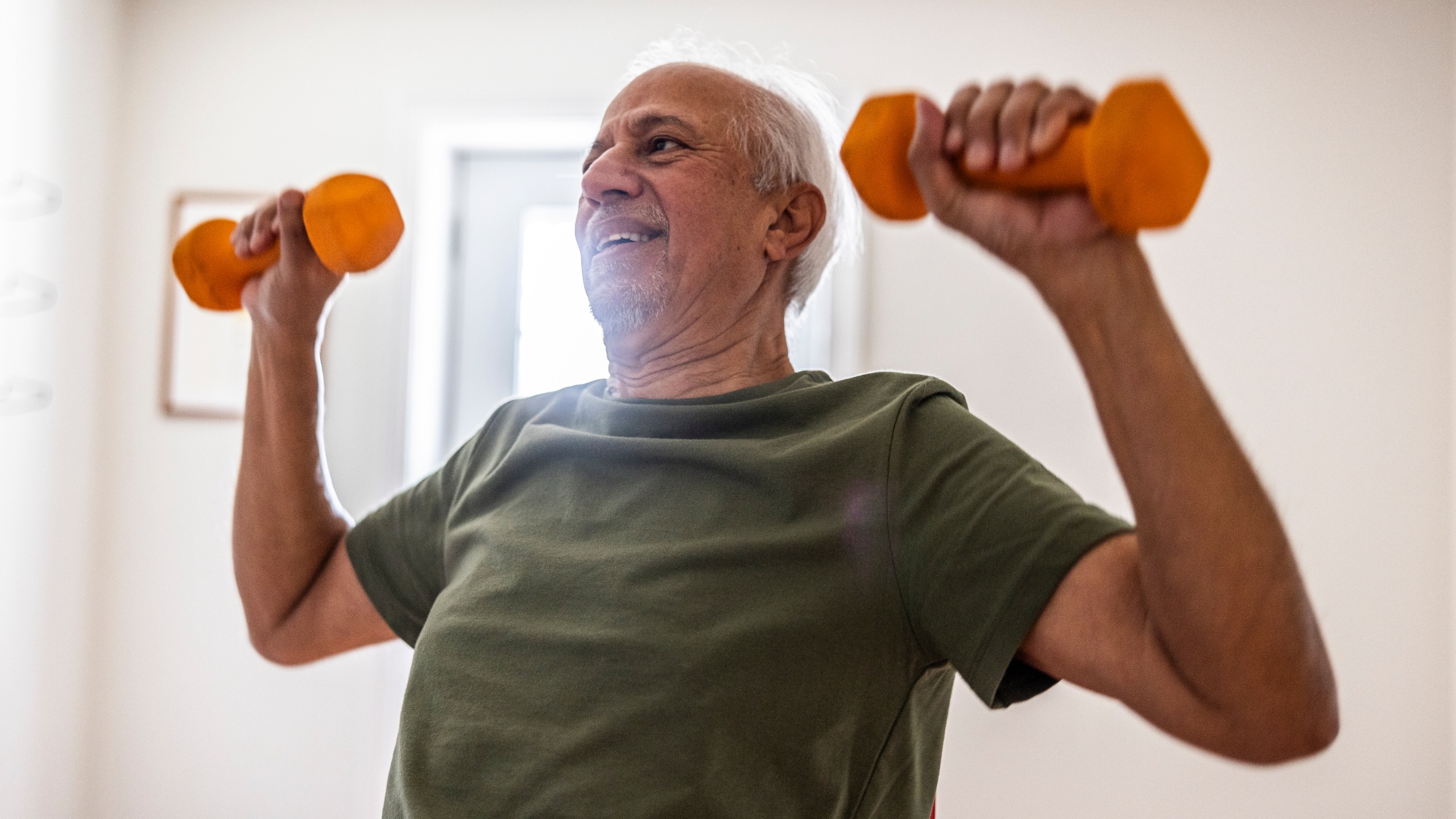 A man in a t-shirt lifts dumbbells in a white room.