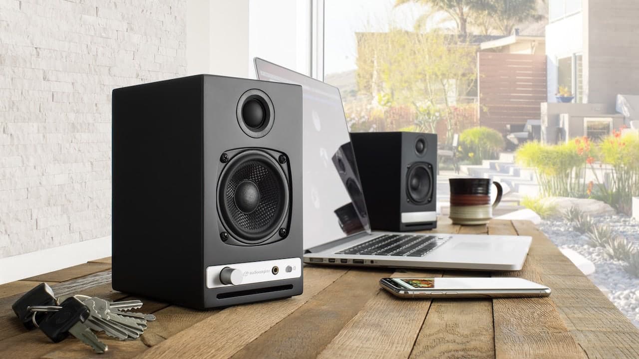 Audioengine HD3 Next-Gen (2025) speakers on a wooden desk next to a phone and laptop and set of keys. There&#039;s a window at the back and you can see a nice garden.