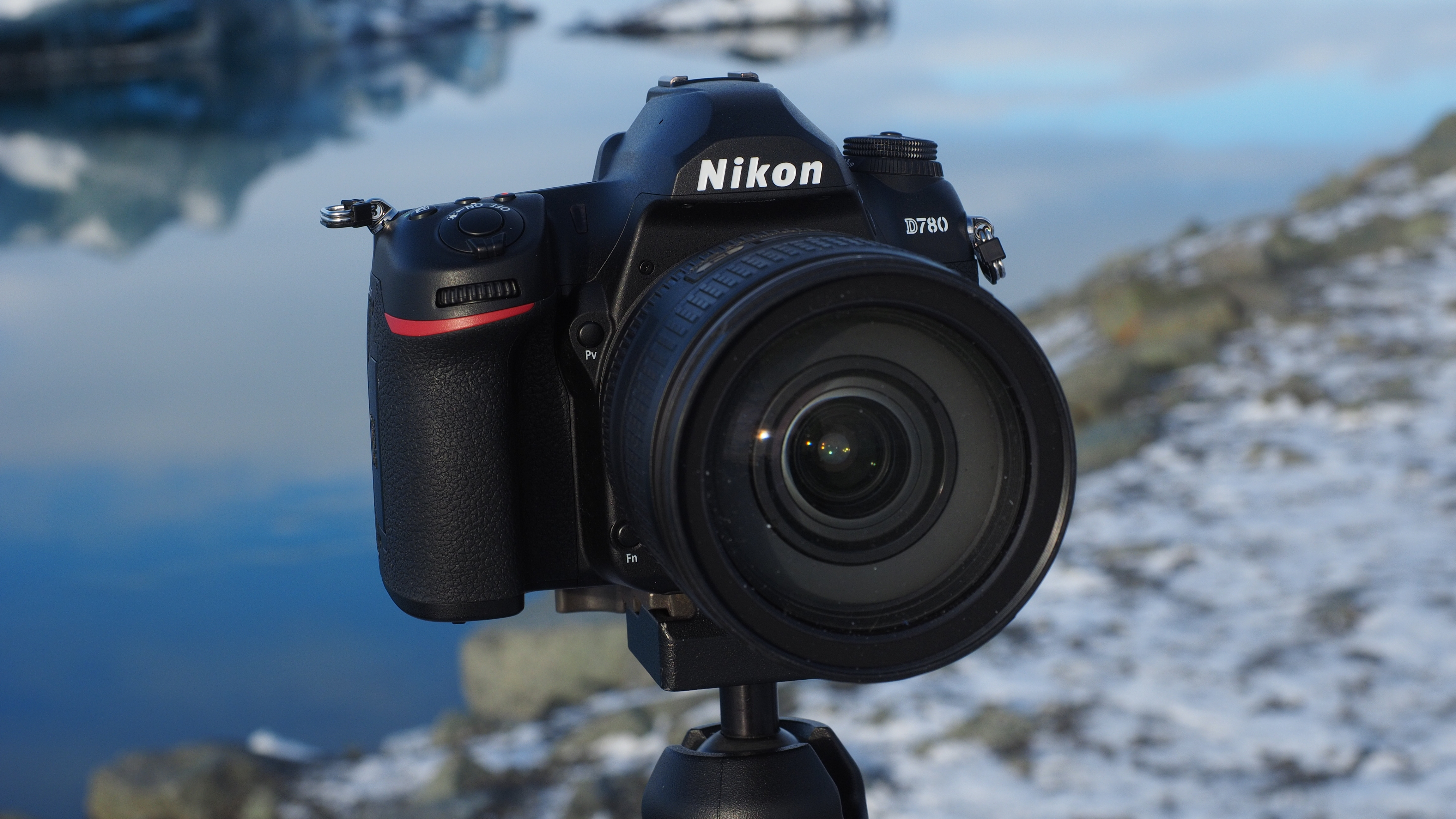 Nikon D780 DSLR camera mounted on a tripod outdoors near a body of water with a rocky shoreline in the background