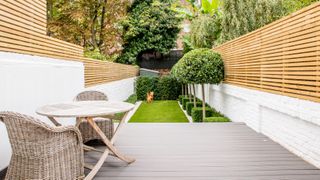 A backyard with decking, brick wall with white paint decor, wooden fencing, a round outdoor table with rattan outdoor chair furniture