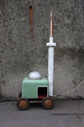 Concrete wall and floor, pastel green piece of square art on four wheels, silver dome on top, grey poll at the back with copper rod top, wire cord
