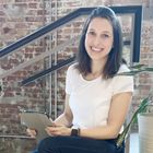 a photo of professional organizer Di Ter Avest sat on a staircase next to a plant: a lady with shoulder length dark hair in a short sleeved white top and navy jeans, holding a notepad