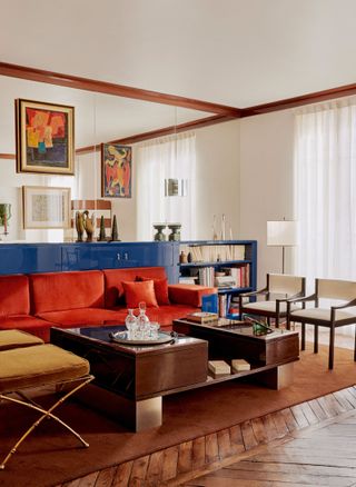 Image of a mid-century modern-style living room with a primary color palette. There are wooden floors and wooden trim around the room. The wall are white, but the back wall is entirely made out of mirrors. There is a low, wall-to-wall cabinet that is royal blue and a red sofa in front of it.