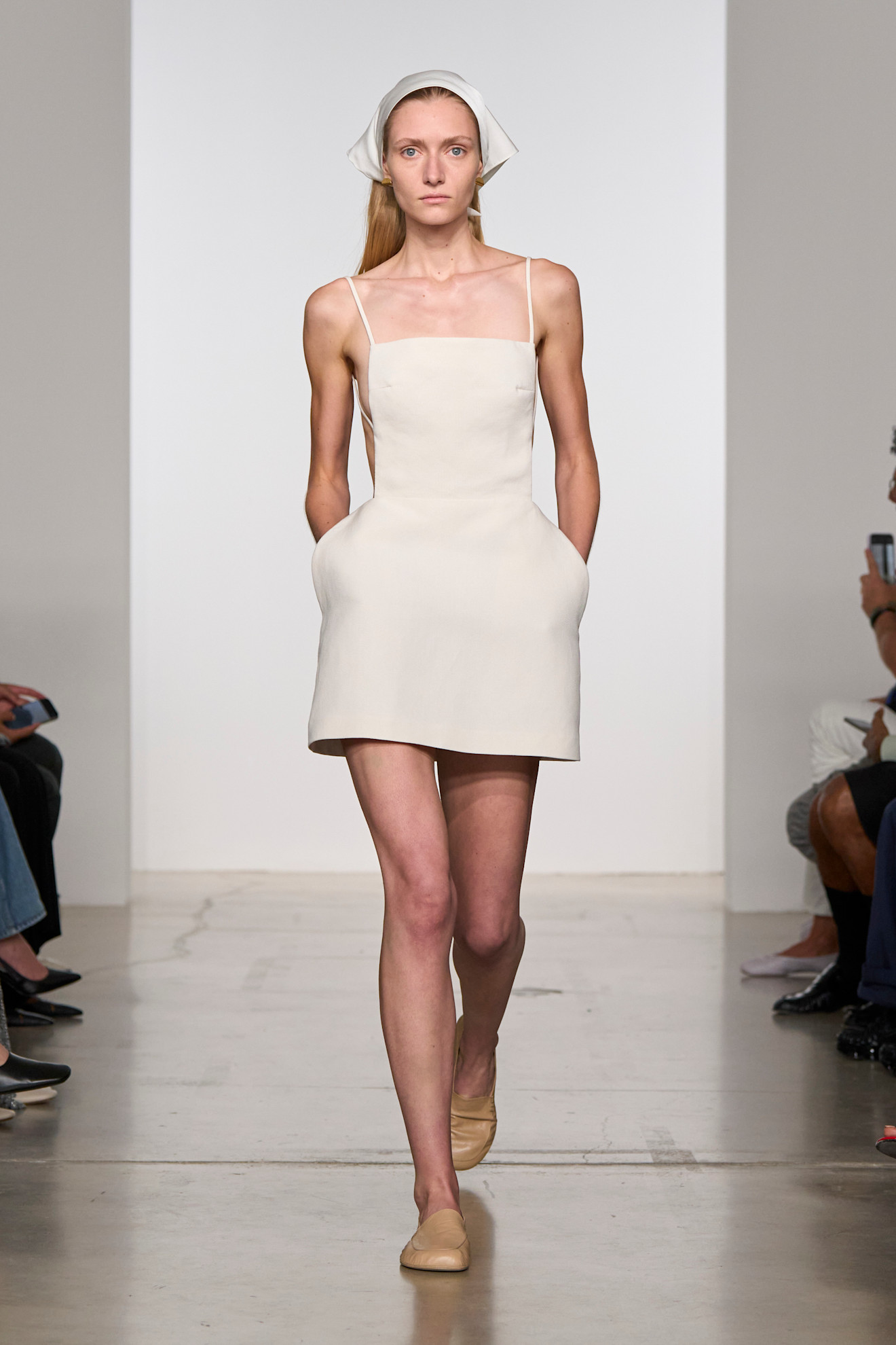 a model walking in Calvin Klein Spring 2026 wearing a white apron dress, white bandana, and tan flats