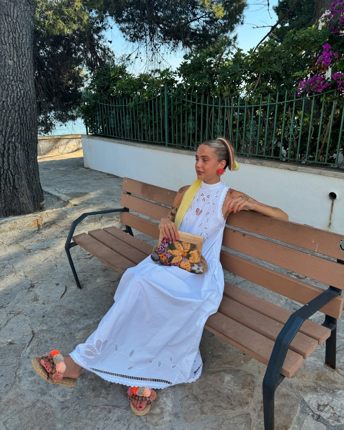 Fashion person Evie Mills wears a long white dress, bobbled sandals and a large multi coloured crochet scrunchie with a long yellow train. She accessories with fluffy earring and a multicolour coloured raffia clutch bag on a recent image from her Instagram.