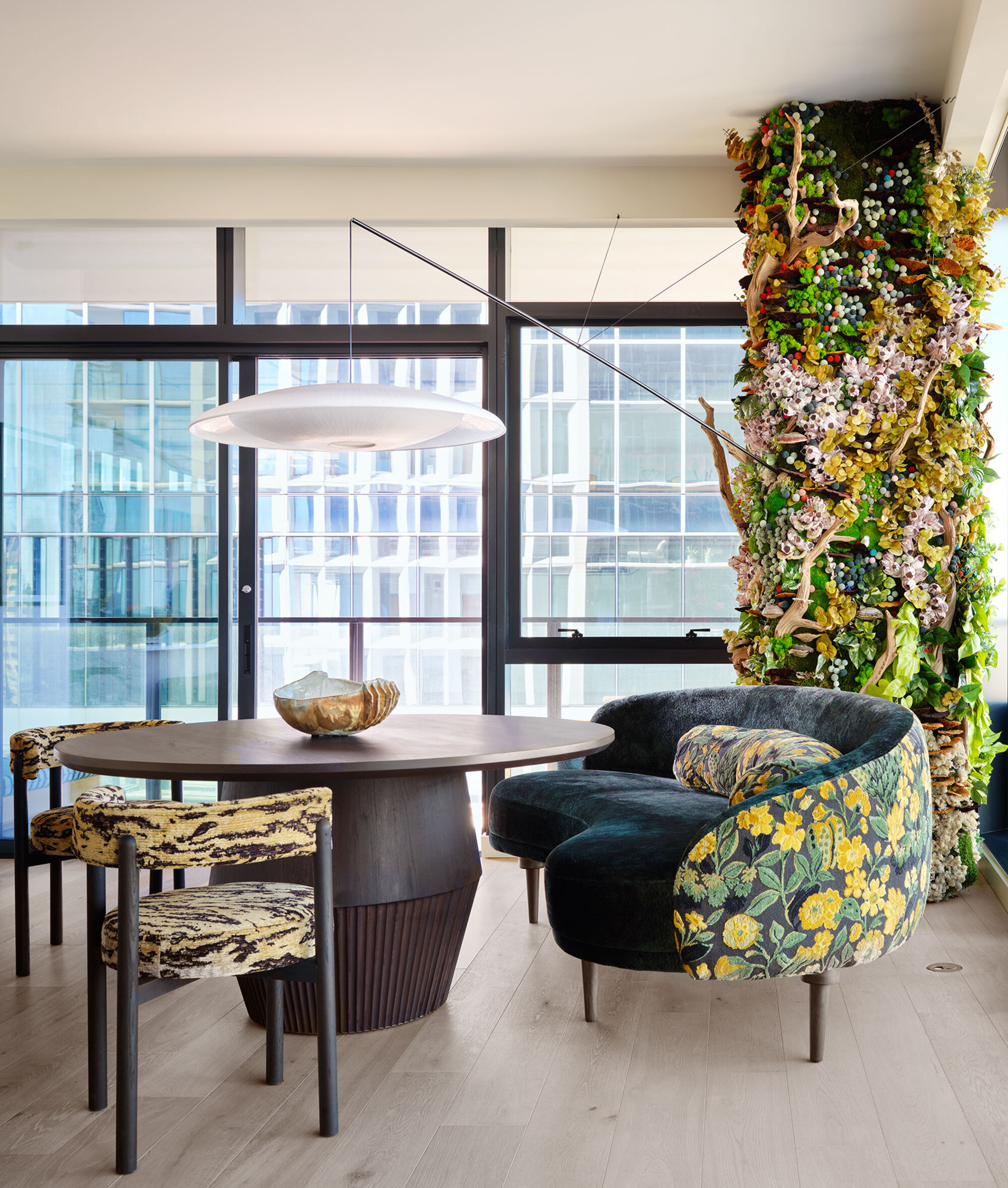 dining room in an apartment with a column covered in a living wall and flowers, next to a round timber coffee table with couch with floral design, and two armchairs, a pendant light hanging over the table, and glass doors