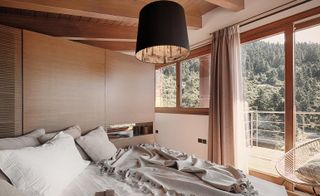 An unlaid bed with 4 pillows with a glass sliding door at the foot of the bed with views of the forest.