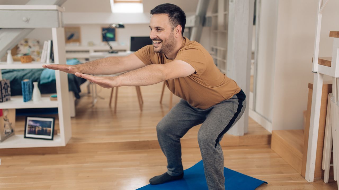 Smiling man performing squat exercise at home, in gray sweatpants and beige T-shirt