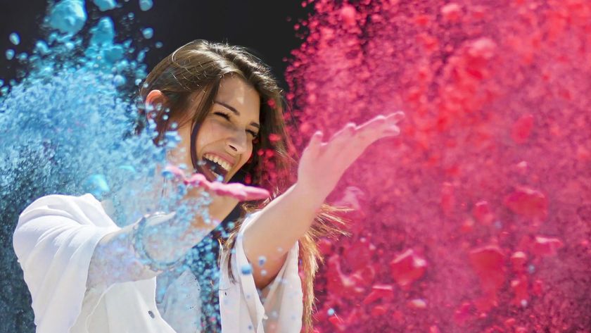 A woman wearing a white shirt against a black background is caught in the moment she's throwing loads of pink and blue powder into the air for a vibrant outdoor portrait
