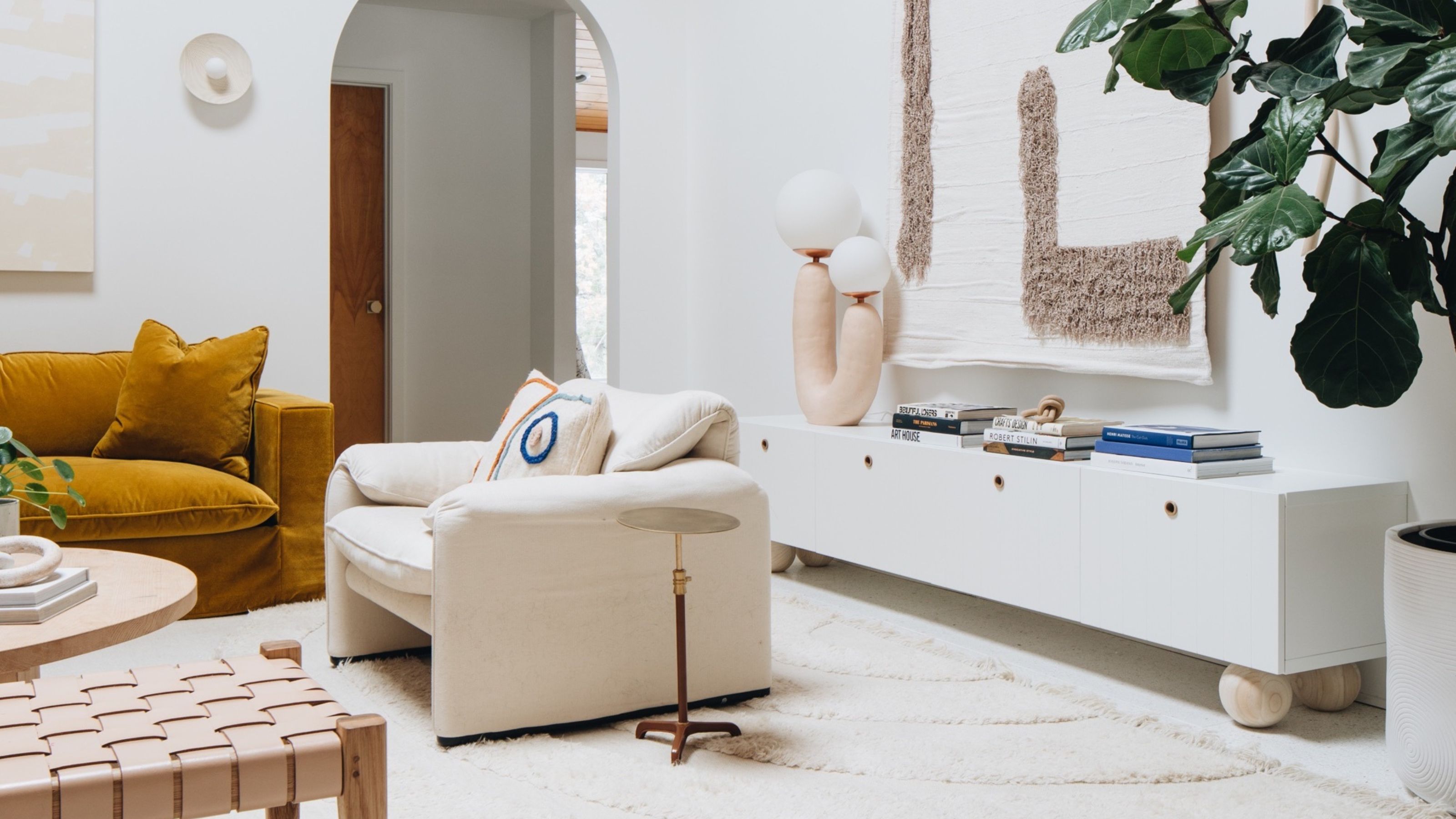 modern boho living room with IKEA besta unit with ball feet added, styled with books and a statement lamp with a a wall hanging above, and potted fiddle leaf beside. elsewhere in the room is a rust-colored velvet sofa, a white armchair with a side table, and a woven bench seat